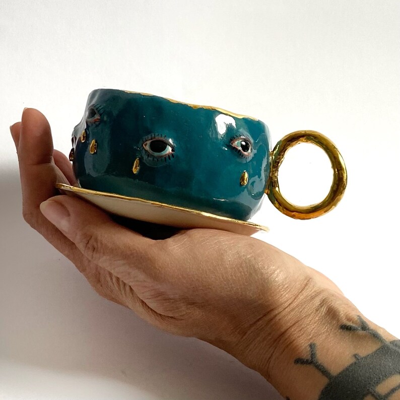 K&ouml;nnte beinhalten: Eine teeblaue Keramik-Teetasse mit goldfarbenem Rand und Henkel. Die Tasse hat drei bemalte Augen mit goldenen Tr&auml;nentropfen, die von jedem Auge tropfen.