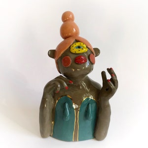May include: A ceramic sculpture of a person with a single large eye, red lips, and a teal blue dress. The sculpture has a peach-colored hair bun and gold accents.