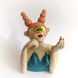 May include: A ceramic sculpture of a figure with a single eye, red lips, and a teal top. The figure has a light skin tone, with pink blush and red nail polish. The hair is peach-coloured with round, ball-like shapes.