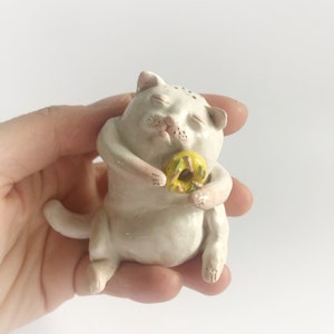 Puede incluir: Figura de cerámica blanca de un gato con los ojos cerrados sosteniendo una dona amarilla con glaseado rosa y chispas verdes.