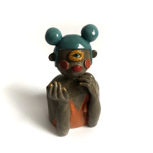 May include: A ceramic figurine of a person with a single large eye, blue hair, and orange clothing. The figurine has a whimsical and colourful design.