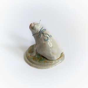 May include: A ceramic pig figurine, seated and looking upwards, with a light blue ribbon. The pig is off-white with pink details, on a round base with green and brown tones. The base is approximately 10 cm in diameter.