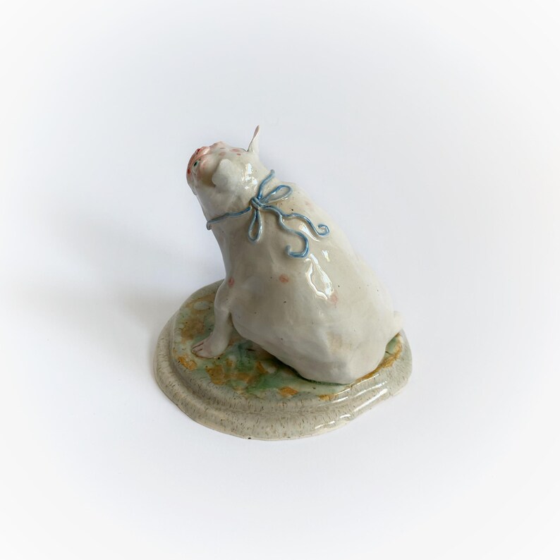 May include: A ceramic pig figurine, seated and looking upwards, with a light blue ribbon. The pig is off-white with pink details, on a round base with green and brown tones. The base is approximately 10 cm in diameter.