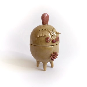 May include: A ceramic trinket box with a whimsical design. It has a round body with a pink top, a face with red eyes, and a pink flower on the side. The box is supported by three legs.
