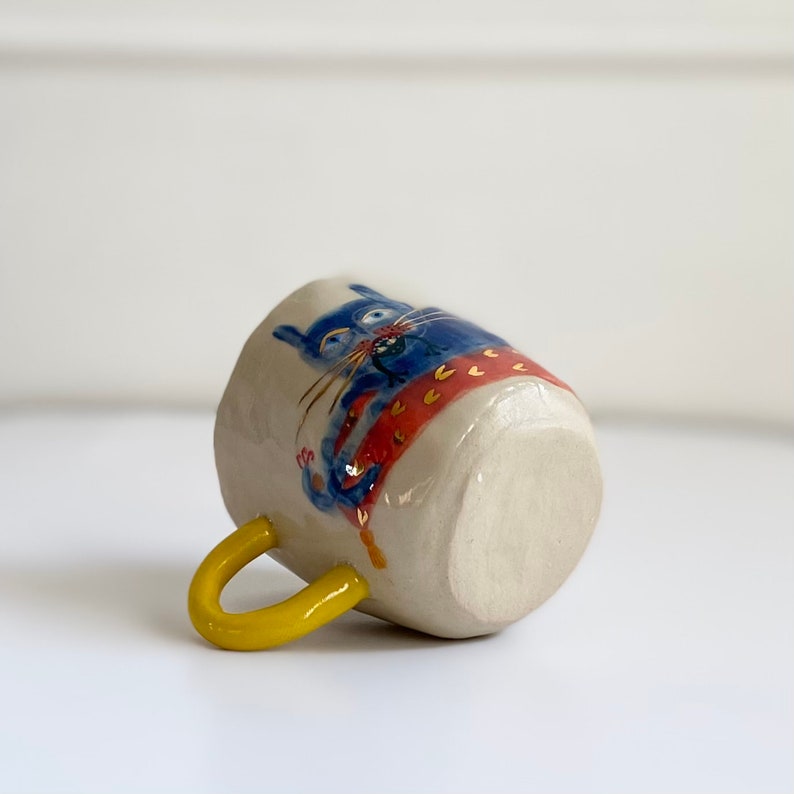 Puede incluir: Una taza de cer&aacute;mica blanca con un asa amarilla. La taza est&aacute; decorada con un gato azul que lleva una bufanda roja y naranja.