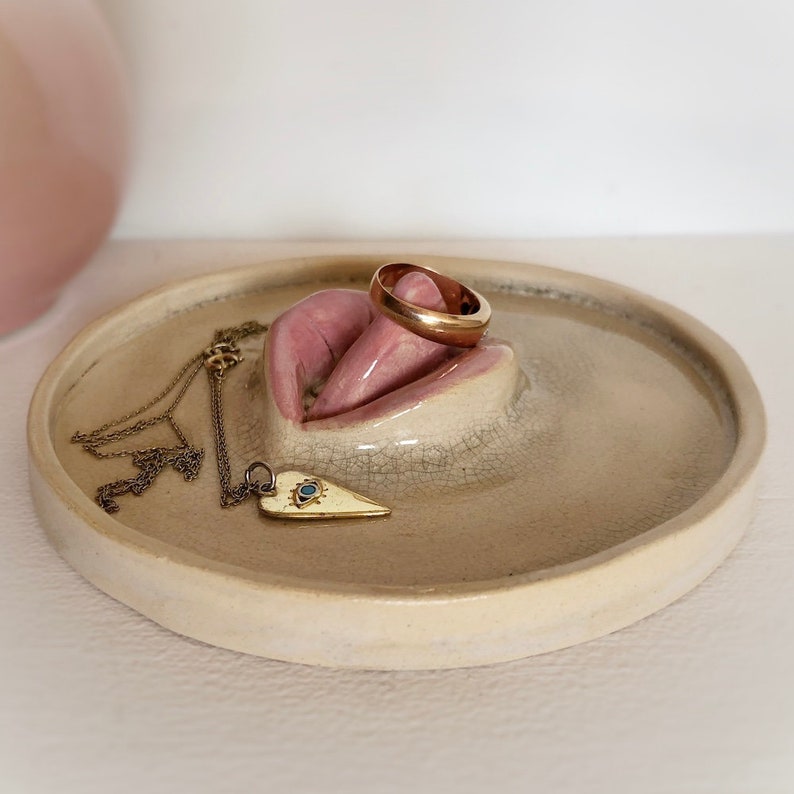 Pode incluir: Um prato de cer&acirc;mica para joias em forma de l&aacute;bios com uma alian&ccedil;a de ouro e um colar de ouro em forma de cora&ccedil;&atilde;o com um pingente de olho mau.