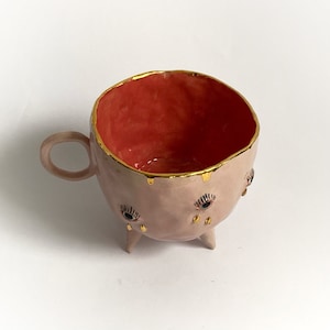 May include: A pink ceramic mug with gold accents and three legs. The mug has a red interior and three eyes with gold eyelashes and black pupils.