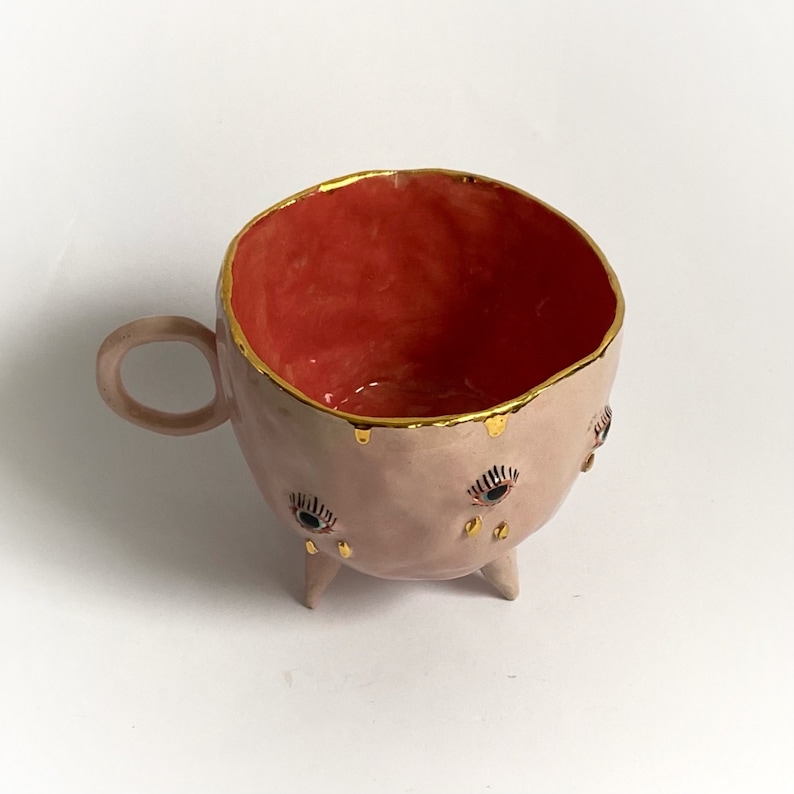May include: A pink ceramic mug with gold accents and three legs. The mug has a red interior and three eyes with gold eyelashes and black pupils.