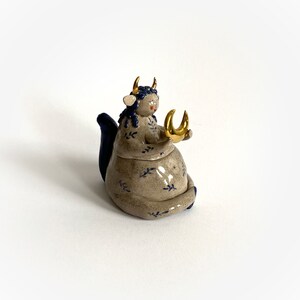 May include: A ceramic figurine of a seated figure with a bovine head, blue hair, and gold horns. The figure wears a gray dress with blue floral patterns and holds a gold crescent moon. The figurine has a blue tail.