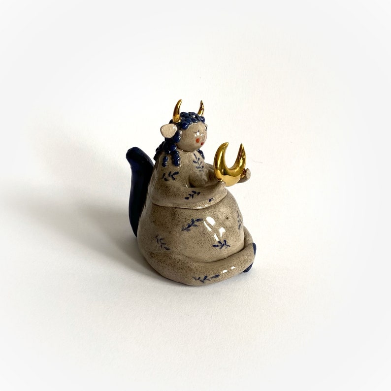 May include: A ceramic figurine of a seated figure with a bovine head, blue hair, and gold horns. The figure wears a gray dress with blue floral patterns and holds a gold crescent moon. The figurine has a blue tail.