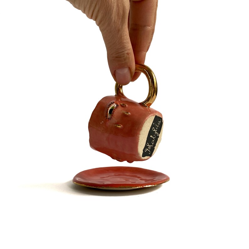 Puede incluir: Una taza de t&eacute; de cer&aacute;mica rosa con asa dorada y un dise&ntilde;o de ojo dorado. La taza tiene una etiqueta negra que dice "Isolatida". La taza est&aacute; sentada sobre un platillo rosa a juego.