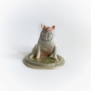 May include: A ceramic sculpture of a white bulldog with pink accents, gazing upwards. The dog sits on a round, green and grey base. A light blue collar is around the dog's neck. The sculpture has a glossy finish.