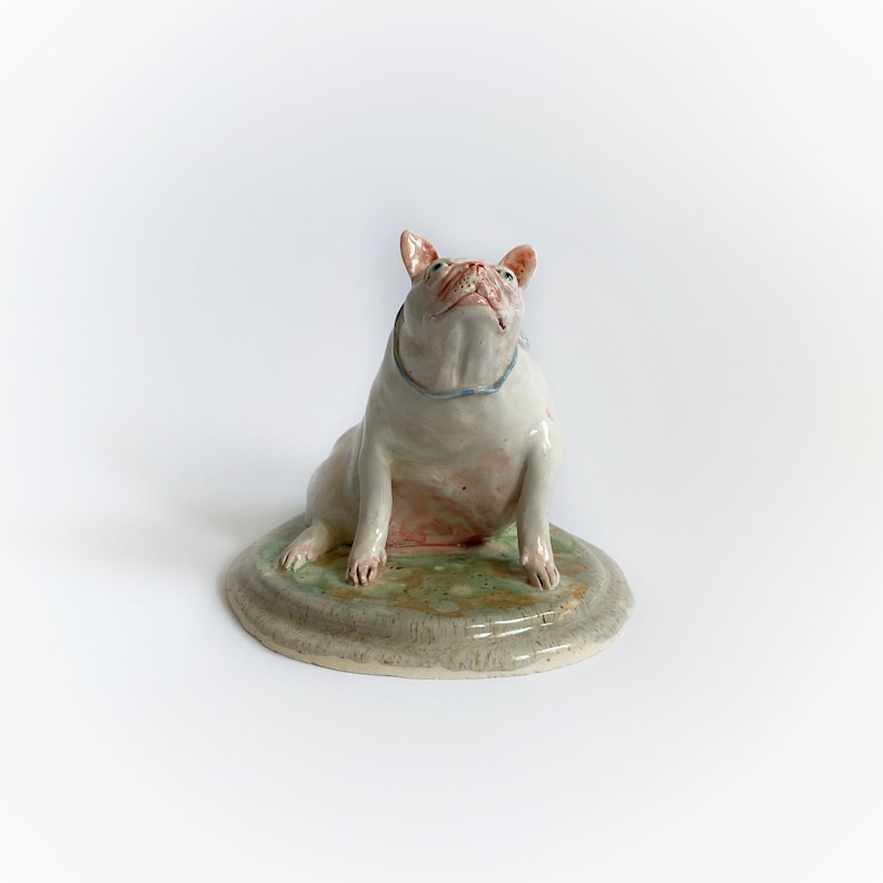 May include: A ceramic sculpture of a white bulldog with pink accents, gazing upwards. The dog sits on a round, green and grey base. A light blue collar is around the dog's neck. The sculpture has a glossy finish.