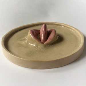 Pode incluir: Um prato de cer&acirc;mica com um design em relevo texturizado de uma boca com uma l&iacute;ngua rosa que se projeta. O prato &eacute; de cor bege claro com um esmalte craquelado.