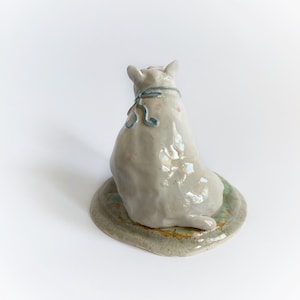 May include: A ceramic cat figurine, viewed from the back, sitting on a round, textured base. The cat is light grey with pink spots and a blue ribbon. The base is a muted green and tan colour.