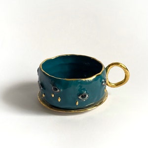 K&ouml;nnte beinhalten: Eine teeblau gef&auml;rbte Keramiktasse mit goldfarbenem Rand und Henkel. Die Tasse ist mit mehreren schwarzen Augen mit goldenen Akzenten und goldenen Tropfenformen verziert.