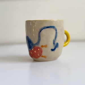 Puede incluir: Una taza de cer&aacute;mica con borde y asa amarillos. La taza est&aacute; decorada con dise&ntilde;os pintados a mano en azul y rojo, incluyendo una flor estilizada con detalles dorados.