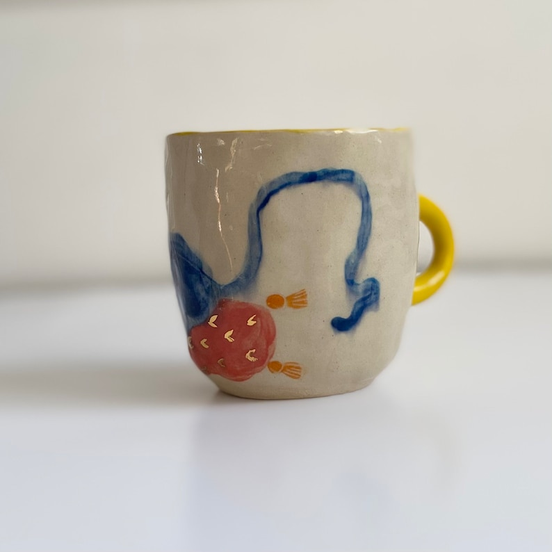 Puede incluir: Una taza de cer&aacute;mica con borde y asa amarillos. La taza est&aacute; decorada con dise&ntilde;os pintados a mano en azul y rojo, incluyendo una flor estilizada con detalles dorados.