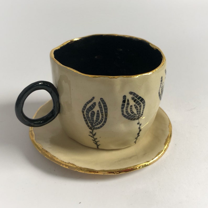 May include: A small, cream-colored teacup and saucer with black floral designs and gold trim.