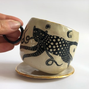 Puede incluir: Una taza de cerámica blanca con una ilustración en blanco y negro de un pulpo con cuerpo manchado. La taza tiene un asa negra y un borde dorado. Está sentada sobre un platillo a juego con un borde dorado.