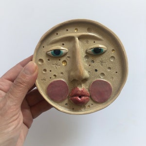 May include: A ceramic plate with a face design. The plate is beige with a cracked texture and features a large nose, red lips, and rosy cheeks. The eyes are blue with gold accents, and there are star and dot details.
