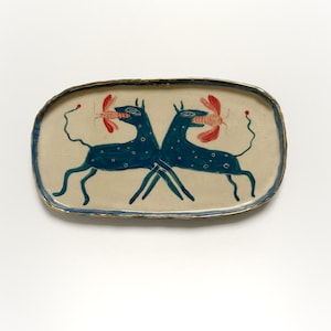 May include: A rectangular ceramic serving tray with a cream base and a blue border. Two teal dog-like figures face each other, each holding a red object. The tray has a gold rim and a whimsical design.