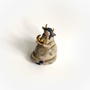 May include: A ceramic figurine of a seated figure with bull horns, blue hair, and a gold crescent moon. The figure is tan with blue floral patterns and accents. The horns and moon are gold. The figurine has closed eyes and a serene expression.