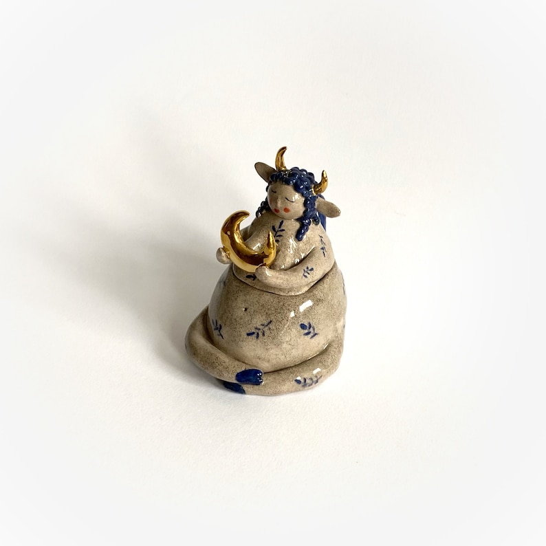 May include: A ceramic figurine of a seated figure with bull horns, blue hair, and a gold crescent moon. The figure is tan with blue floral patterns and accents. The horns and moon are gold. The figurine has closed eyes and a serene expression.