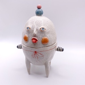 May include: A white ceramic egg-shaped container with a face, red lips, and blue eyes. The container has a lid with a blue and pink ball on top. The container has three legs and black and white striped arms.