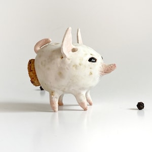 Puede incluir: Una figurita de cerdo de cer&aacute;mica blanca con una cola de corcho y ojos negros. El cerdo est&aacute; de pie sobre una superficie blanca.