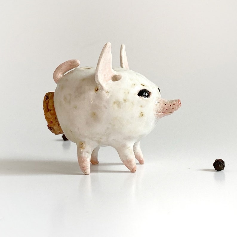 Puede incluir: Una figurita de cerdo de cer&aacute;mica blanca con una cola de corcho y ojos negros. El cerdo est&aacute; de pie sobre una superficie blanca.