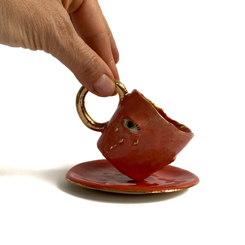 Puede incluir: Una peque&ntilde;a taza de t&eacute; de cer&aacute;mica roja con un borde dorado y un dise&ntilde;o de ojo pintado. La taza est&aacute; inclinada sobre un platillo a juego.