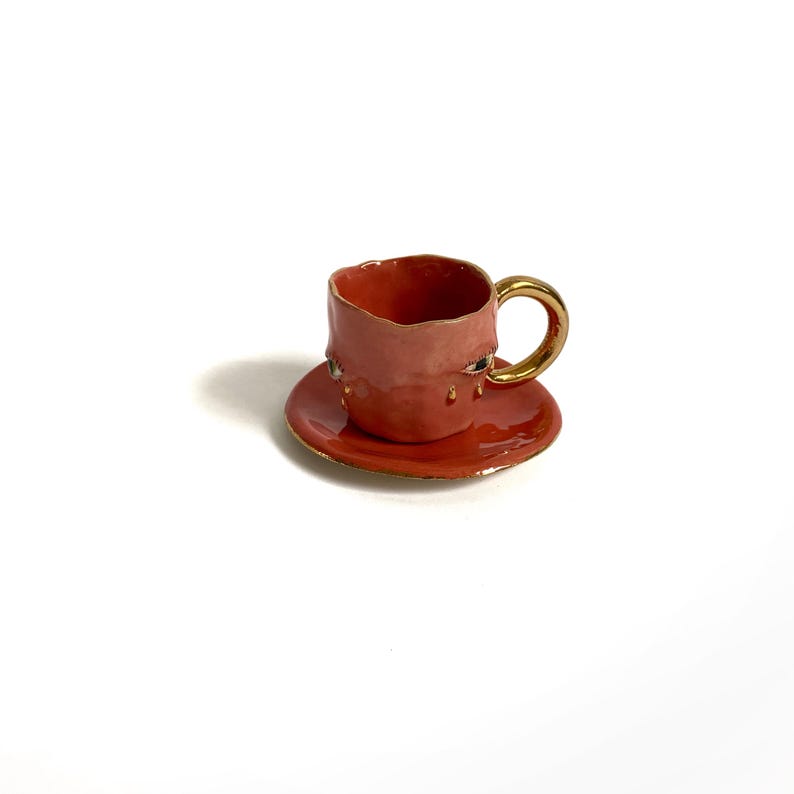 Puede incluir: Una peque&ntilde;a taza de t&eacute; de cer&aacute;mica de color coral con un asa dorada y un platillo a juego. La taza y el platillo tienen una superficie texturizada, ligeramente irregular.