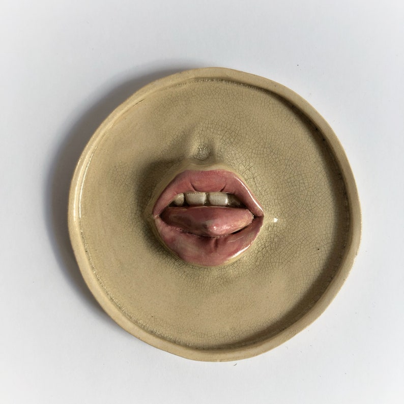 Pode incluir: Uma decora&ccedil;&atilde;o de parede de cer&acirc;mica em forma de prato redondo com uma imagem em relevo tridimensional de uma boca com a l&iacute;ngua de fora.