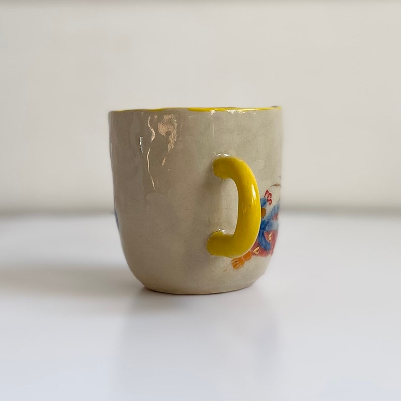 Puede incluir: Una taza peque&ntilde;a de cer&aacute;mica blanca con un asa amarilla y un dise&ntilde;o pintado de colores. La taza tiene un borde ligeramente irregular y un fondo redondeado.