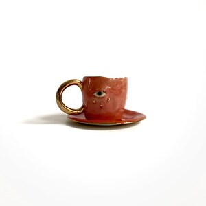 Puede incluir: Una taza de t&eacute; y un platillo de cer&aacute;mica rosa con un borde dorado. La taza de t&eacute; tiene un solo ojo con un iris verde y pesta&ntilde;as doradas pintadas en la parte delantera. El ojo est&aacute; llorando l&aacute;grimas doradas.