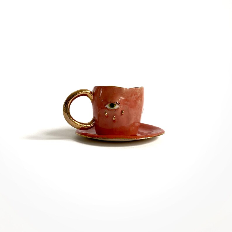 Puede incluir: Una taza de t&eacute; y un platillo de cer&aacute;mica rosa con un borde dorado. La taza de t&eacute; tiene un solo ojo con un iris verde y pesta&ntilde;as doradas pintadas en la parte delantera. El ojo est&aacute; llorando l&aacute;grimas doradas.