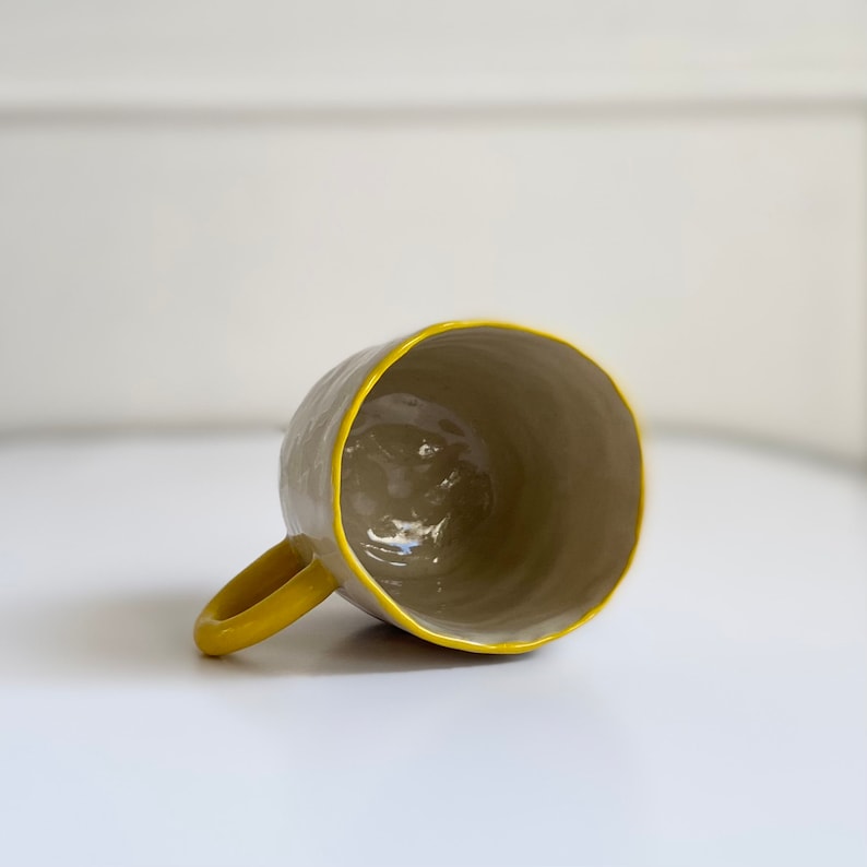 Puede incluir: Una taza de cer&aacute;mica gris con un borde y un asa amarillos. La taza est&aacute; vac&iacute;a y se encuentra sobre una superficie blanca.