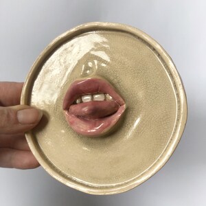 Pode incluir: Uma decora&ccedil;&atilde;o de parede de cer&acirc;mica em forma de boca com uma l&iacute;ngua rosa que se projeta. A boca est&aacute; aberta e tem dentes brancos. O fundo &eacute; de cor bege claro.