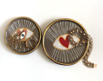 May include: Two ceramic dishes with gold trim, each featuring a stylized eye design. One dish has a blue eye with pink accents, the other has a red heart in the eye. Both dishes have gold accents and are perfect for holding jewelry or other small items.