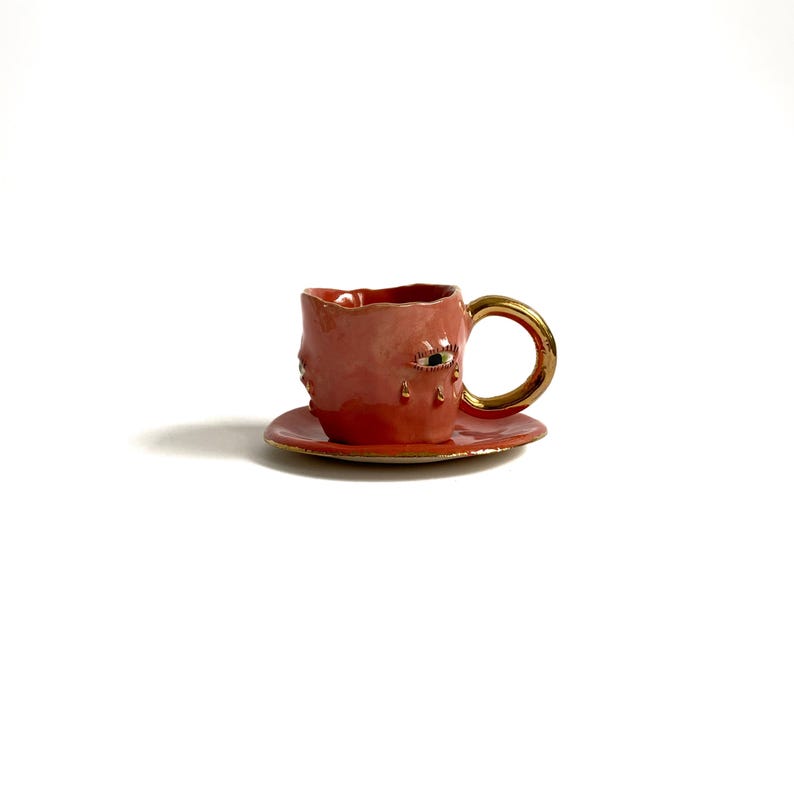 Puede incluir: Una taza de t&eacute; de cer&aacute;mica rosa con un asa dorada y un dise&ntilde;o de ojo &uacute;nico. La taza est&aacute; sentada sobre un platillo a juego.