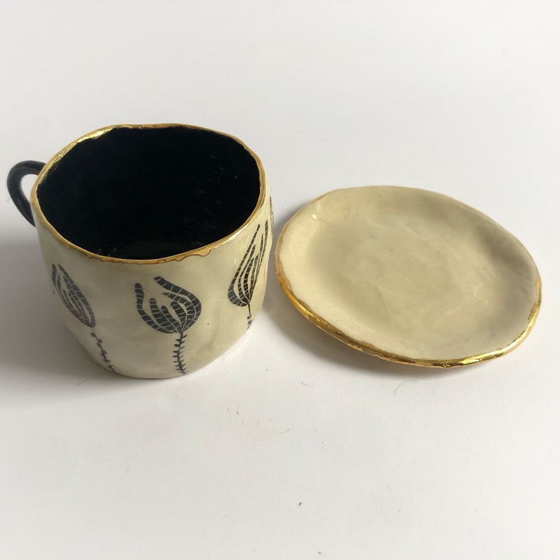 May include: A white ceramic teacup and saucer set with black floral designs and gold accents. The cup has a black interior and a small handle.