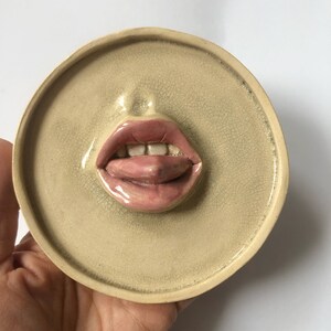 Pode incluir: Uma decora&ccedil;&atilde;o de parede de cer&acirc;mica em forma de boca com a l&iacute;ngua de fora. A boca &eacute; pintada em tons de rosa e branco, e a l&iacute;ngua &eacute; de um rosa mais escuro. O fundo &eacute; bege claro.