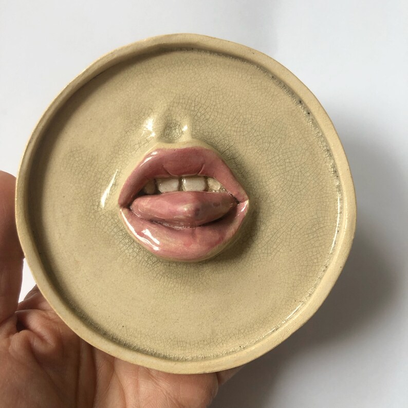 Pode incluir: Uma decora&ccedil;&atilde;o de parede de cer&acirc;mica em forma de boca com a l&iacute;ngua de fora. A boca &eacute; pintada em tons de rosa e branco, e a l&iacute;ngua &eacute; de um rosa mais escuro. O fundo &eacute; bege claro.
