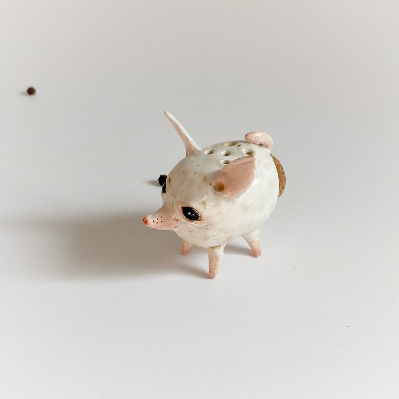 Puede incluir: Una figurita de cerdo de cer&aacute;mica blanca con orejas rosas y ojos negros. El cerdo tiene agujeros en la espalda y est&aacute; de pie sobre cuatro patas. Es un objeto decorativo.