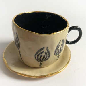 May include: A white ceramic teacup and saucer set with black floral designs and gold accents. The cup has a black handle.