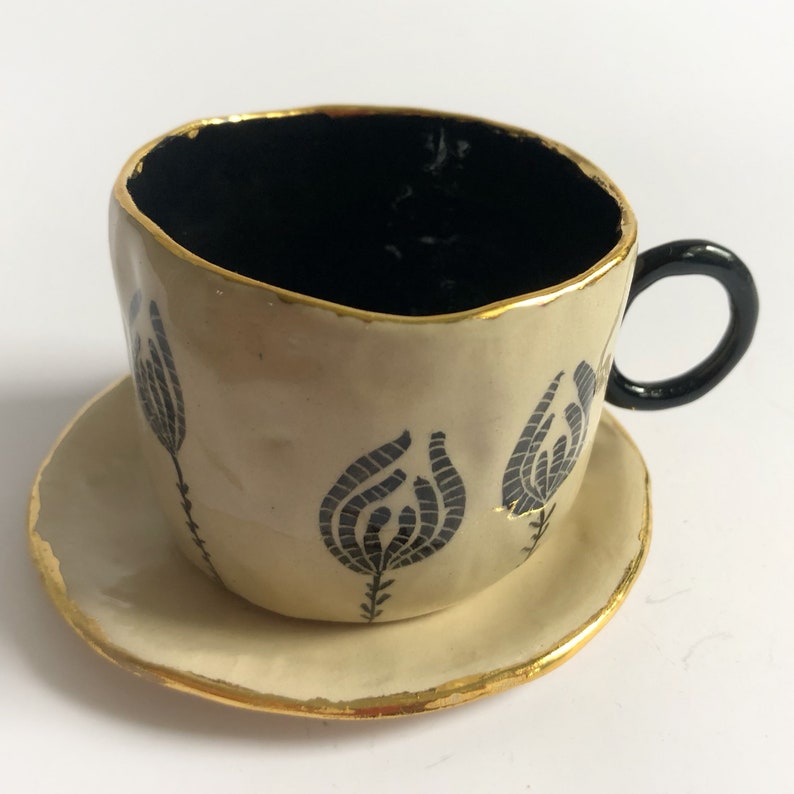May include: A white ceramic teacup and saucer set with black floral designs and gold accents. The cup has a black handle.