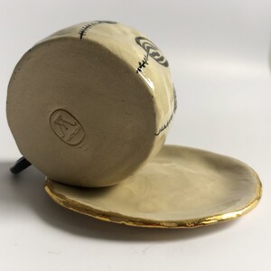 May include: A ceramic mug with a black and white design of stitches and circles. The mug is sitting on a saucer with a gold rim. The bottom of the mug has a maker's mark that reads "AMY LDA".