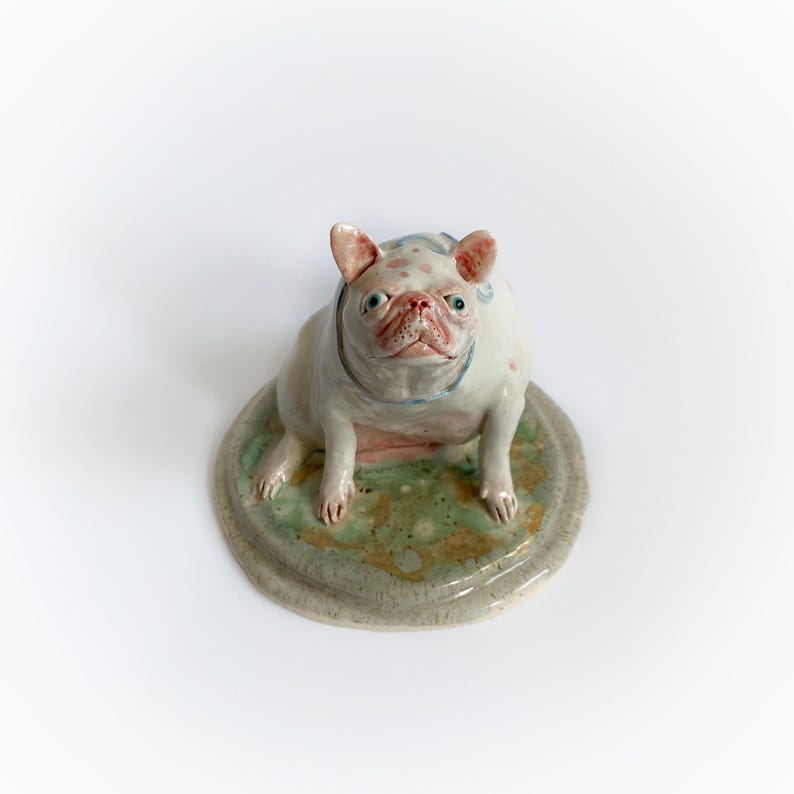 May include: A ceramic bulldog figurine with a white body, pink spots, and a blue collar. The dog sits on a round, green and brown base. The sculpture has a whimsical, artistic style.