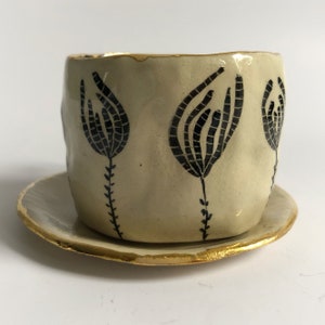 May include: A white ceramic teacup and saucer set with a black floral design and gold trim.